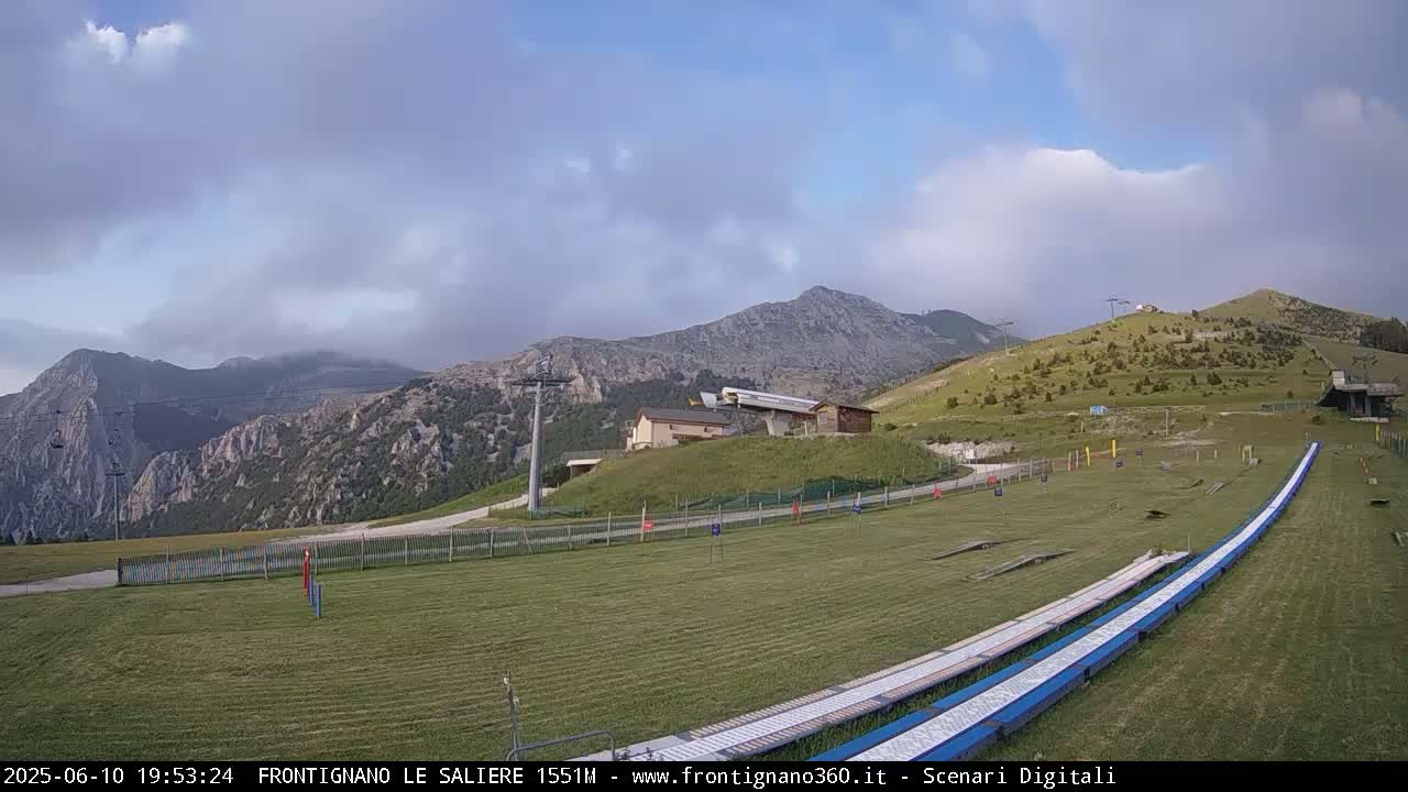 A grassy mountaintop area with a ski lift, small buildings, and two long, blue slides is visible under a partly cloudy sky.