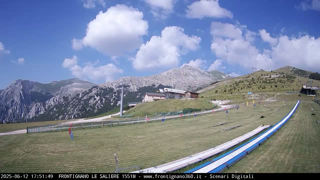 Two long, white slides sit on a grassy area overlooking a mountain range under a partly cloudy sky.