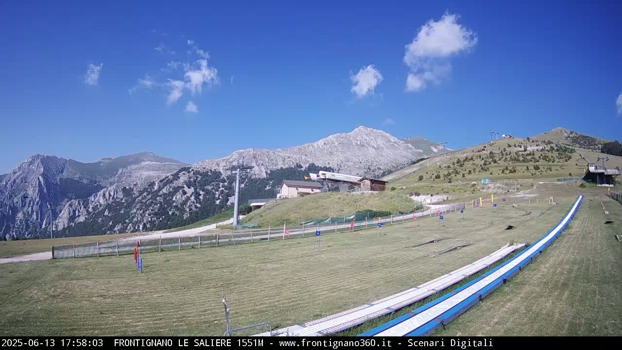 A grassy mountaintop area with a ski lift and long, blue and white slides, under a mostly sunny sky with a few clouds.