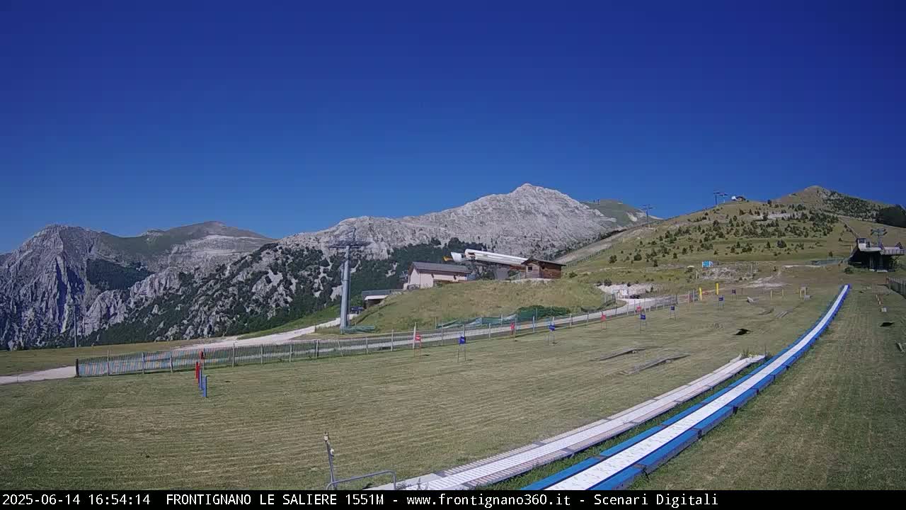 A grassy mountaintop area with a ski lift, small buildings, and long, blue-and-white slides, all under a clear blue sky.