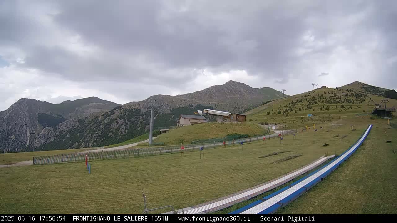 A grassy mountaintop area with a ski lift and two long, blue, downhill slides is visible under an overcast sky.