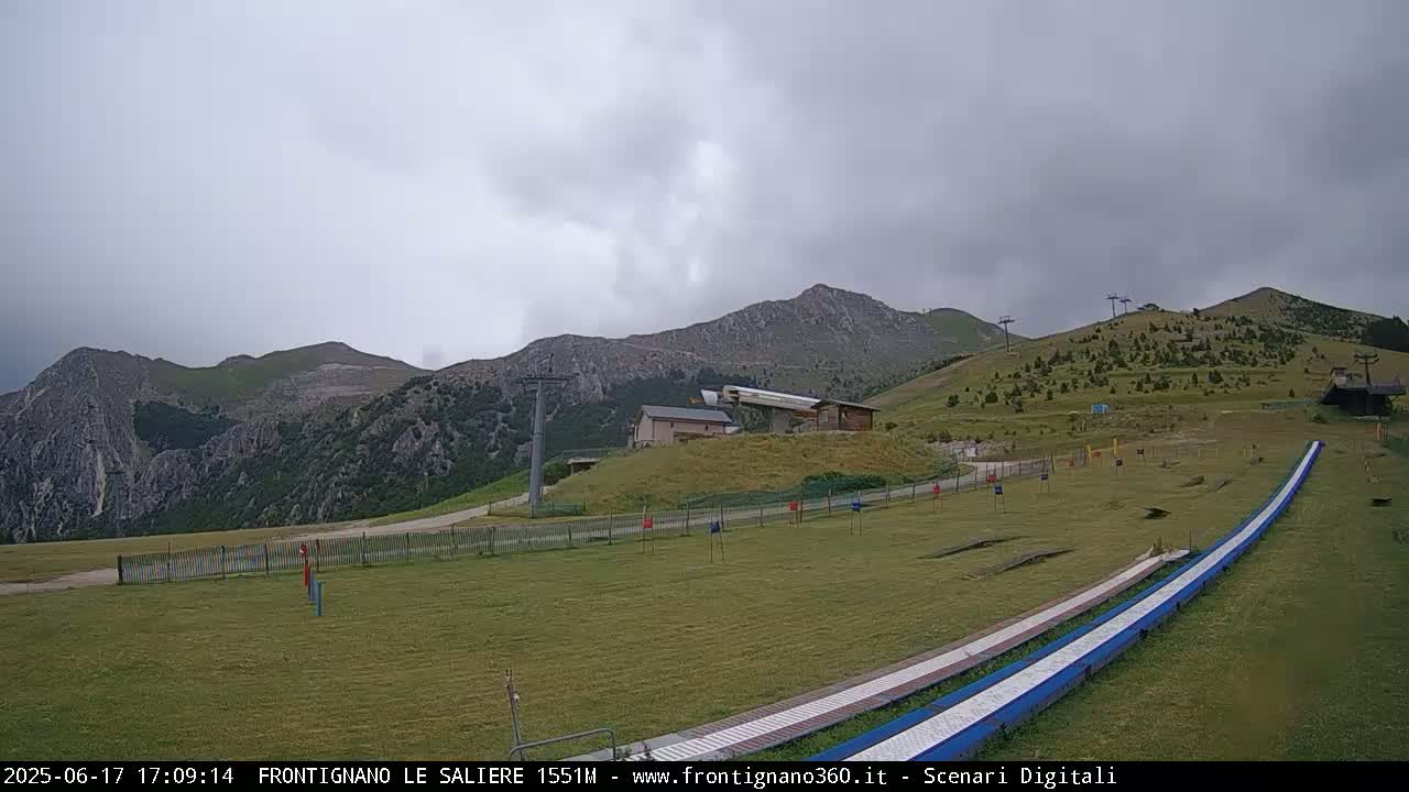 A grassy mountainside with a ski lift, a small building, and two long, flat slides under an overcast sky.