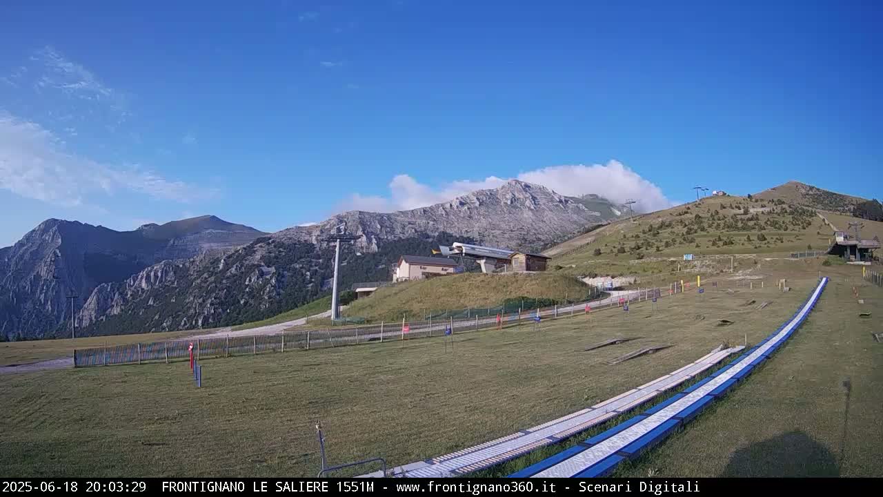 A grassy mountainside with a ski lift and a few buildings, under a mostly clear blue sky with some clouds.
