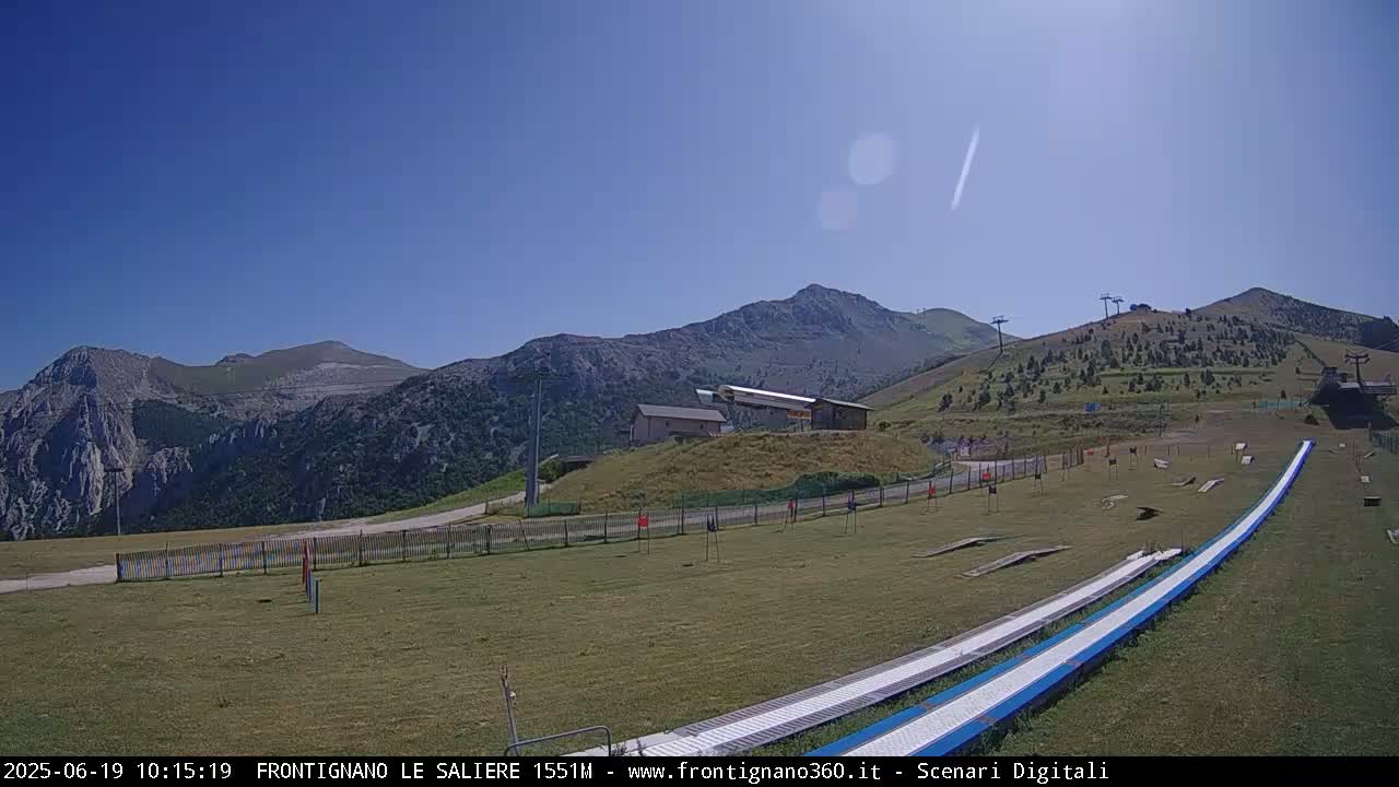 A grassy mountainside with a ski lift, a building, and a long, blue slide, under a clear blue sky.