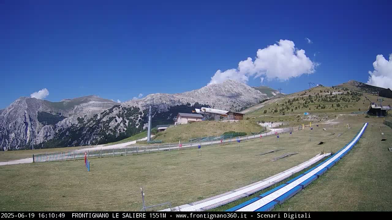 A grassy mountaintop area with a pair of long, blue slides, a few buildings, and a chairlift in the background under a mostly sunny sky with some fluffy clouds.