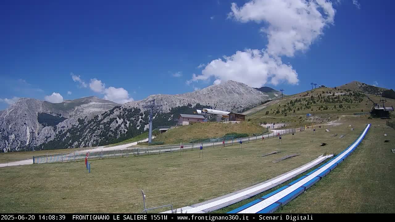 A mountaintop grassy area with a ski lift and two long, flat, light-blue ramps under a mostly sunny sky with some fluffy white clouds.