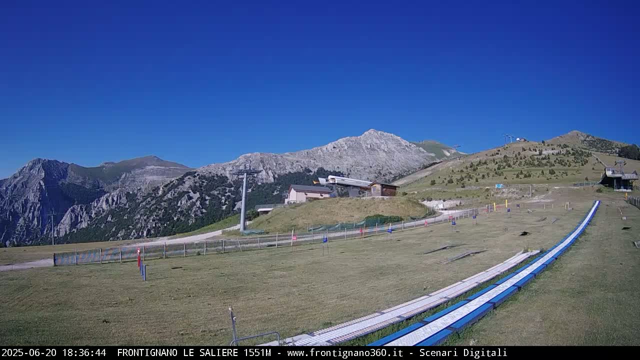 A grassy mountaintop area with a ski lift and several structures, under a clear blue sky.