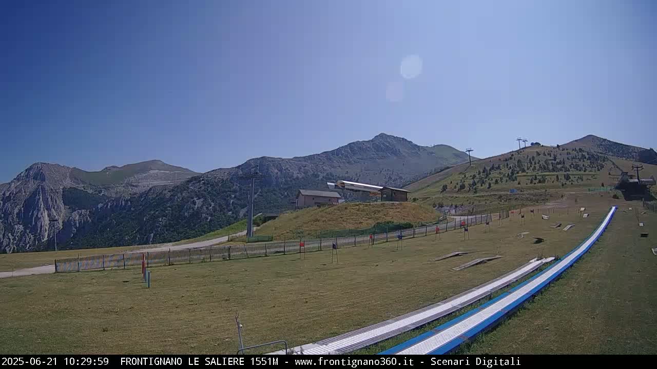 A grassy mountainside area with ski lifts, a small building, and two long, light-blue slides stretches out under a clear blue sky.