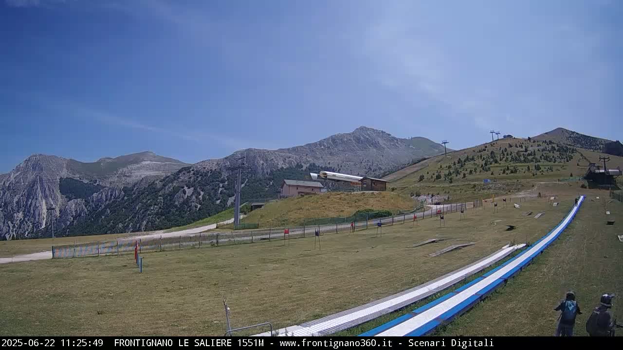 A sunny mountaintop view shows a grassy area with two long, low, blue and white ramps, a small building, ski lifts, and mountains in the background.