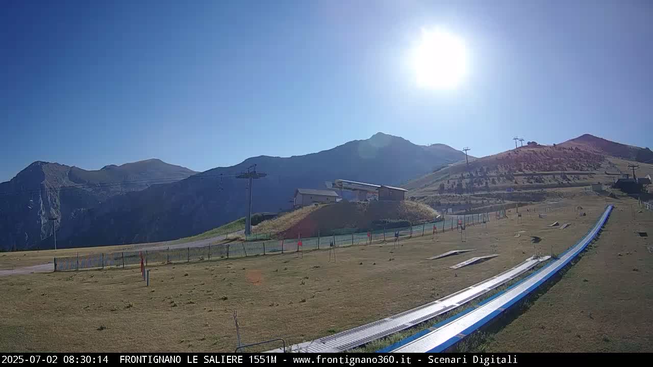 A sunny day reveals a mountain landscape with a long, flat, blue slide in the foreground and ski lifts in the distance.