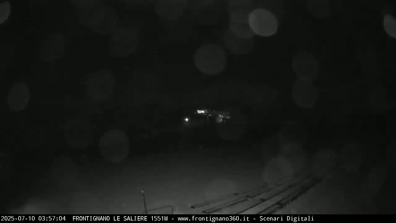 A nighttime, blurry view of a dark area with faint lights in the distance, possibly rain or snow falling.