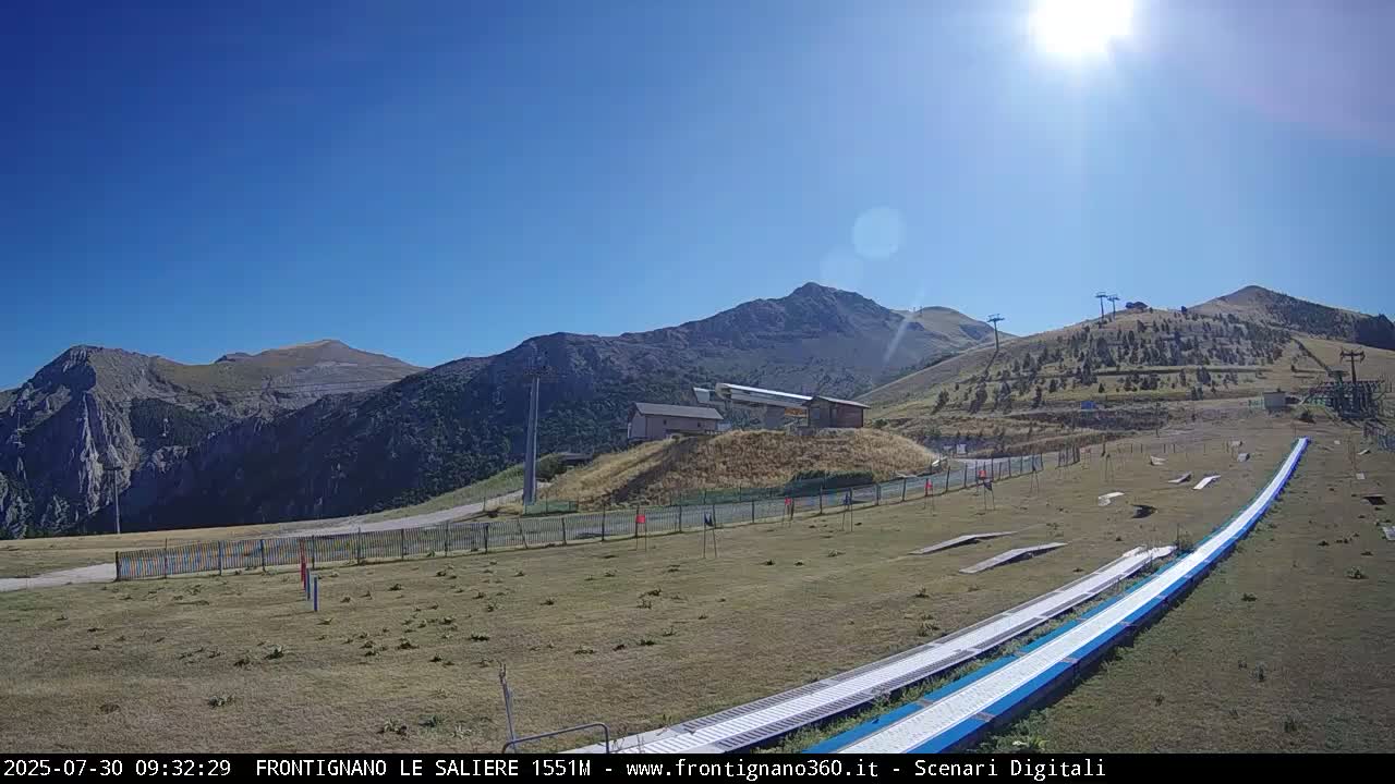 A sunny day at a mountaintop ski resort shows a long, snowless, summer luge track leading towards a ski lift and several mountain peaks in the background.