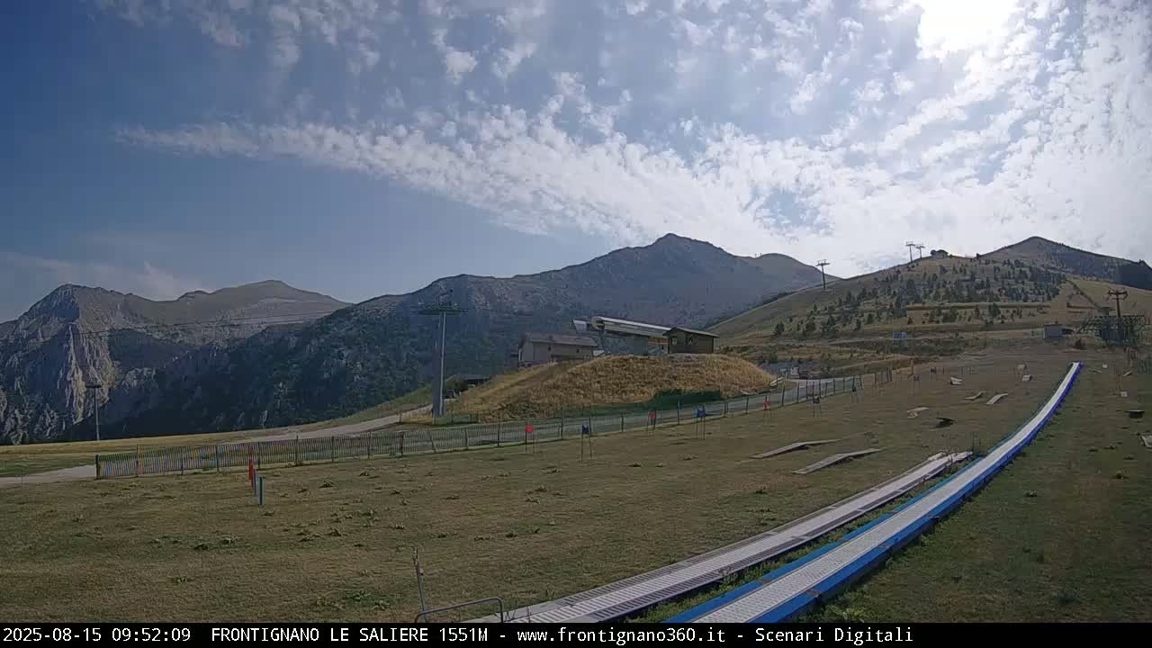 A mostly sunny day with some clouds displays a mountain landscape featuring ski lifts, a grassy area with a long, low structure, and rolling hills in the background.