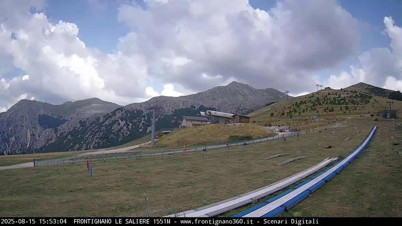 A mostly sunny day with some clouds displays a mountain landscape featuring ski lifts, a grassy area with a long, low structure, and rolling hills in the background.