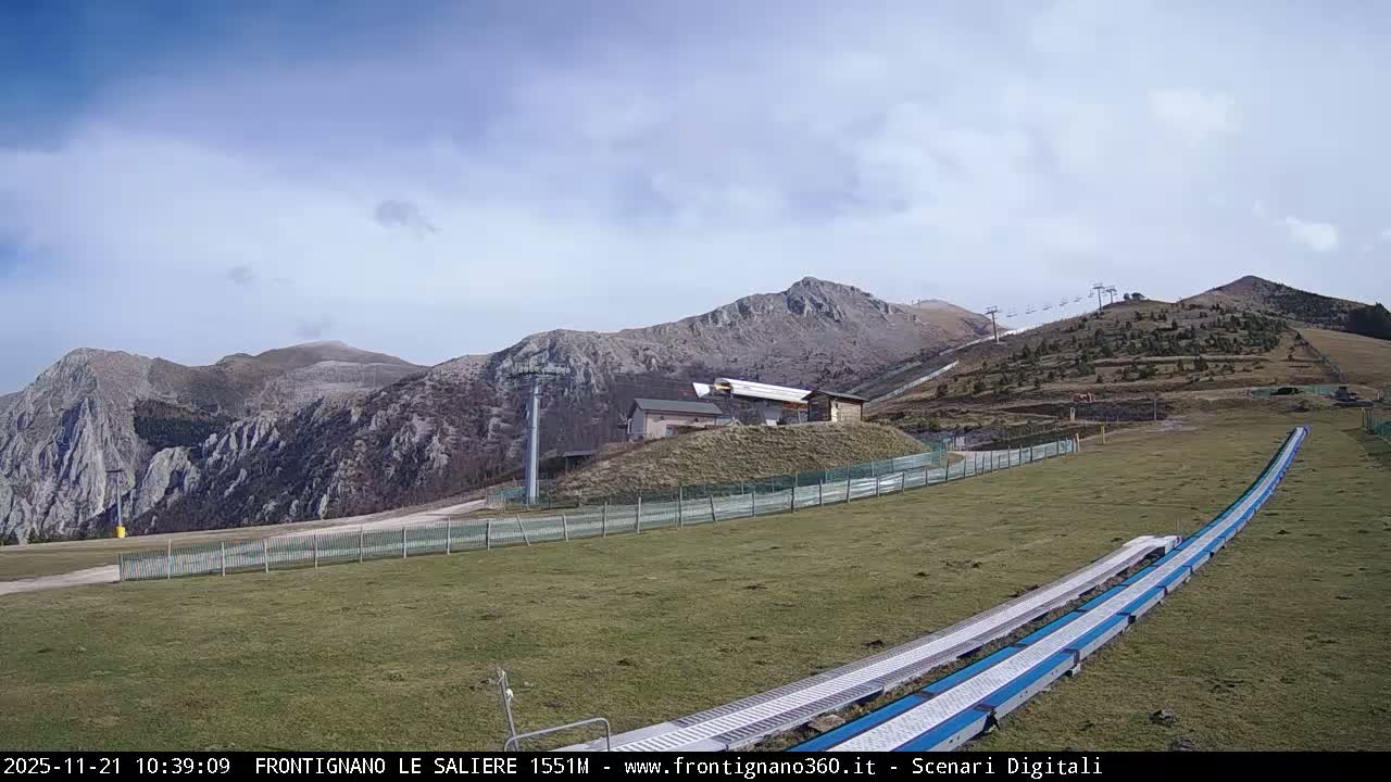 Under a partly cloudy sky, a mountain landscape features green grassy slopes with a bobsled track in the foreground, rocky peaks, and ski lift pylons leading towards buildings nestled on a hill in the midground.