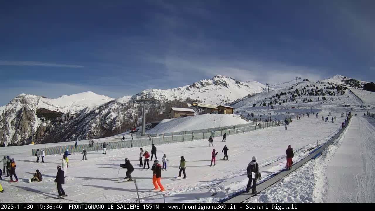 On a bright, sunny day with a clear blue sky, a busy ski resort features numerous skiers and snowboarders on its snow-covered slopes, set against a backdrop of towering, snow-dusted mountains and operational ski lifts.