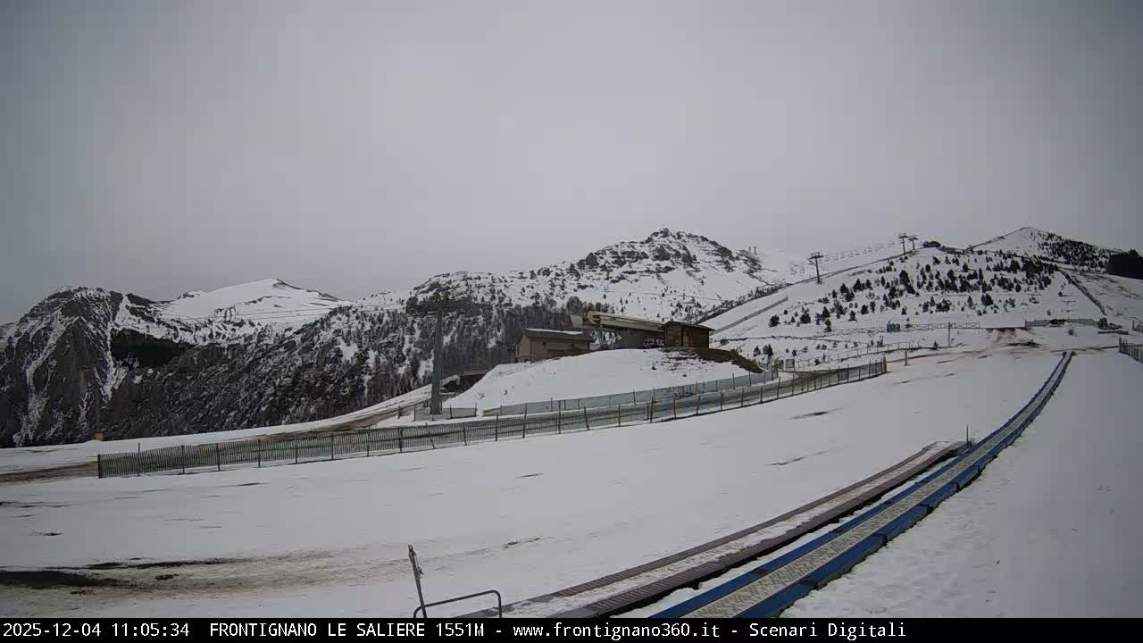 A vast snow-covered mountain landscape featuring ski slopes, several buildings, and numerous ski lifts is visible under a uniformly overcast and grey sky.