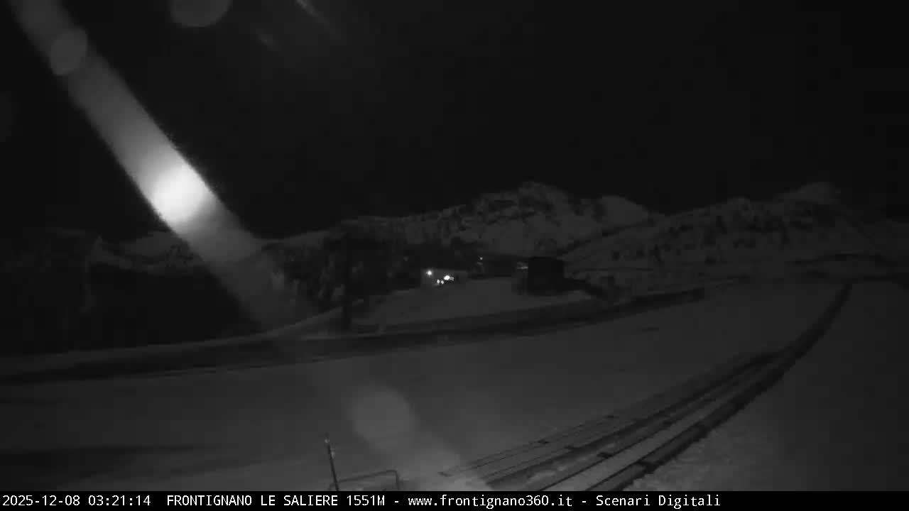 A monochromatic nighttime view depicts a snow-covered mountain landscape with distant illuminated buildings and a bright, diagonal spotlight cutting through the dark sky on a clear, cold night.