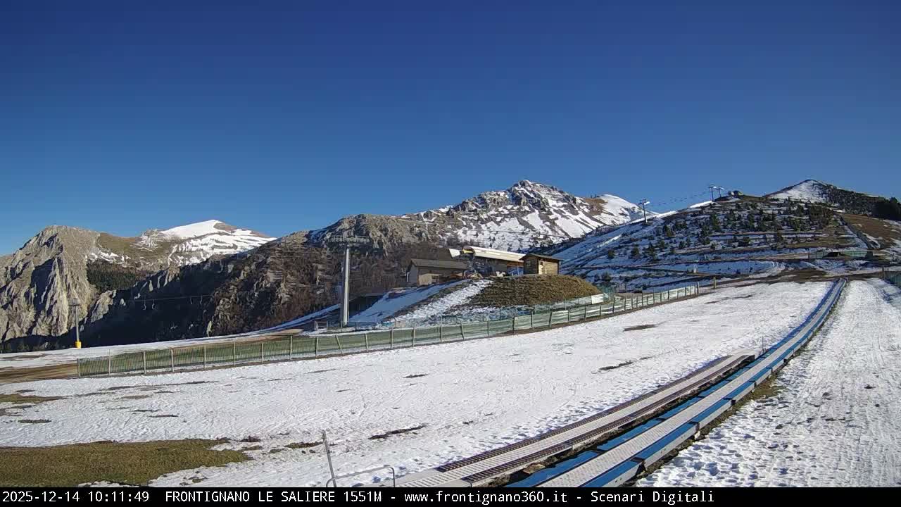A monochromatic nighttime view depicts a snow-covered mountain landscape with distant illuminated buildings and a bright, diagonal spotlight cutting through the dark sky on a clear, cold night.