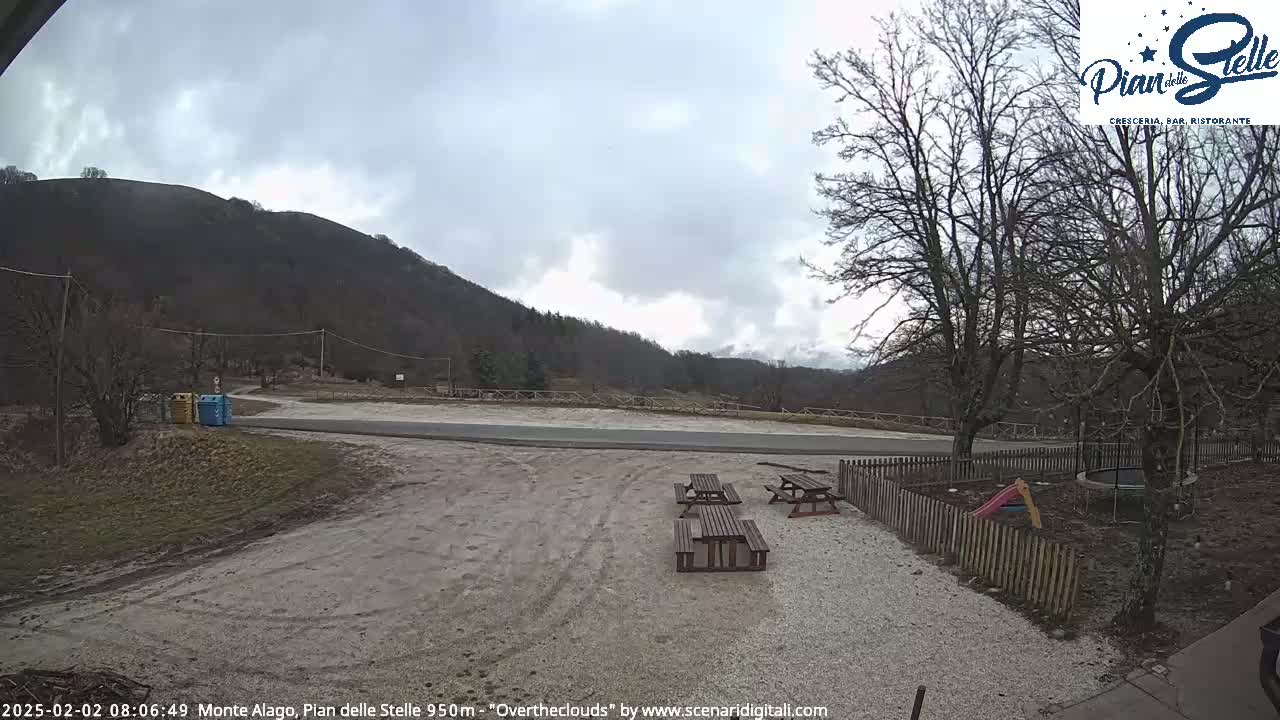 A gravel area with picnic tables sits in front of a wooden fence, overlooking a paved road and a mountain under an overcast sky.