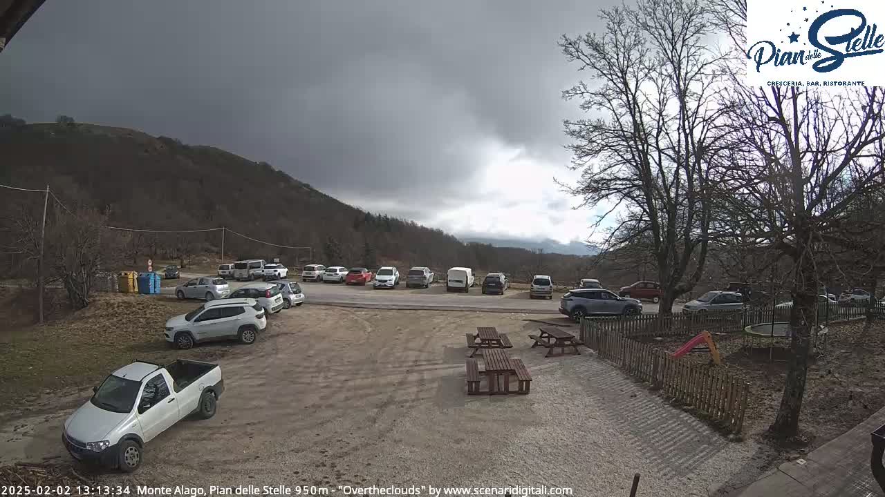 A parking lot with numerous cars and a pickup truck is situated near several picnic tables and a small playground, all set against a backdrop of a mountain under an overcast sky.