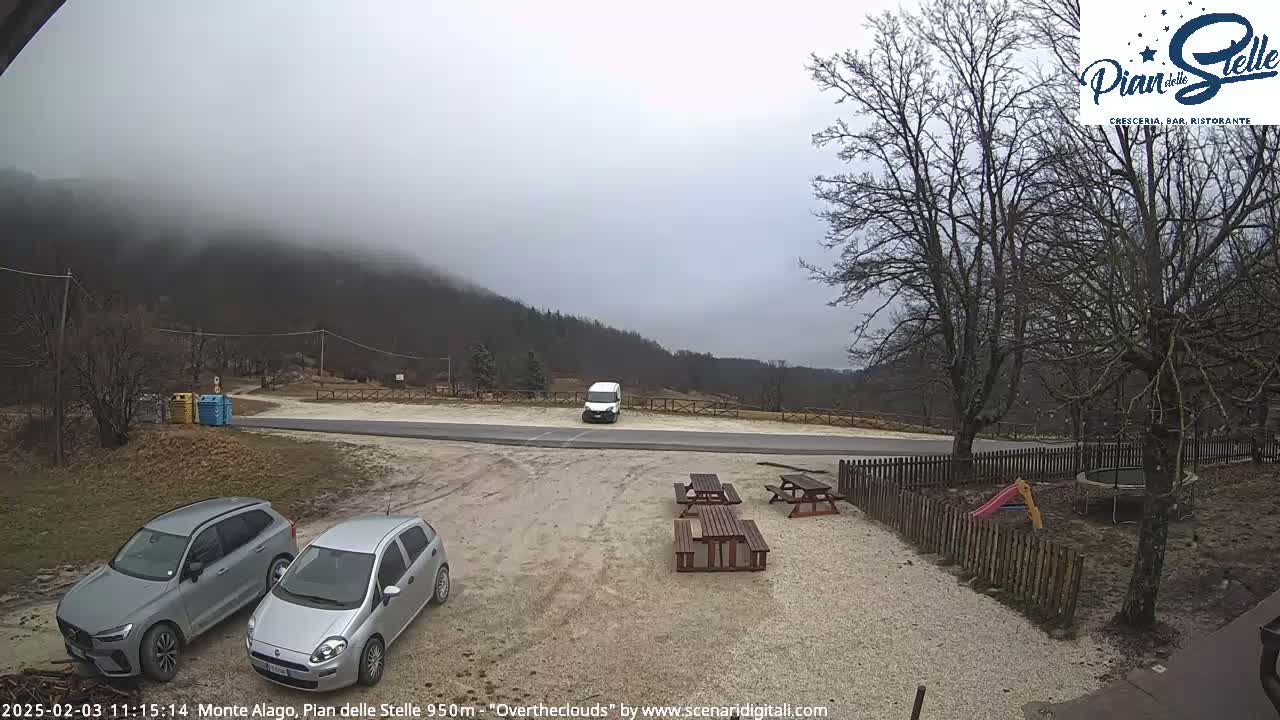 A parking lot with two cars and picnic tables is situated in front of a mountain shrouded in fog, with leafless trees and a small playground visible nearby.