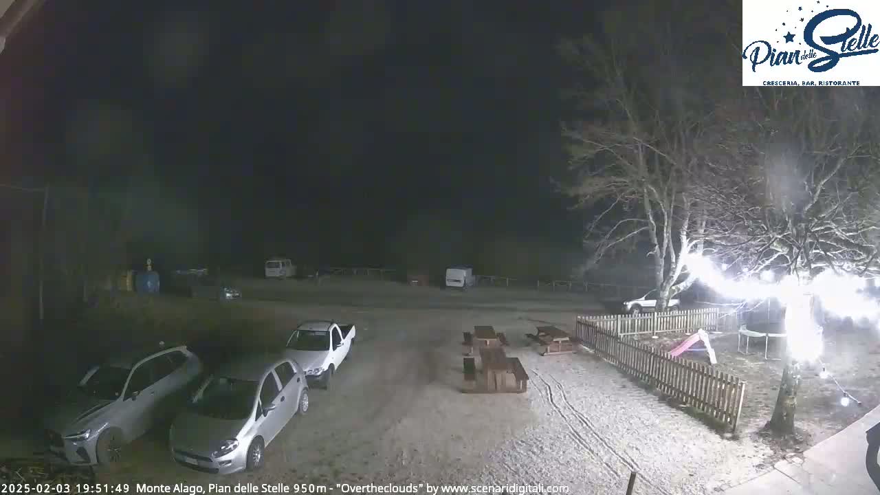 A snow-covered parking lot at night, with several cars parked, picnic tables, and a small playground visible near a tree with string lights.