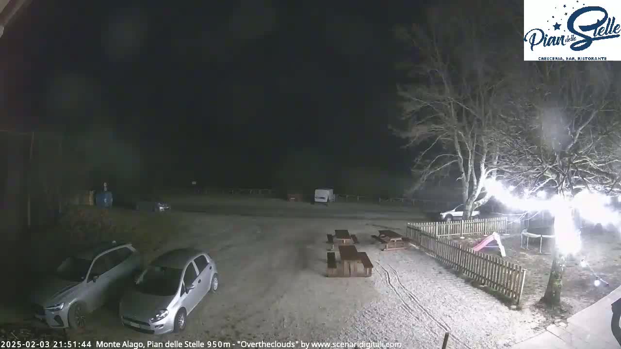 A snow-covered parking lot at night, with several cars parked, picnic tables, a playground, and a tree with bright lights strung around it.