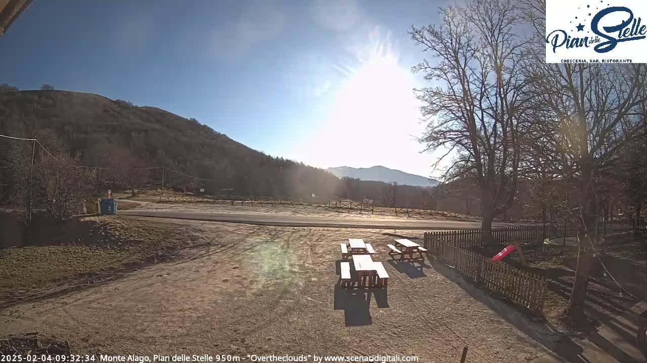 A sunny day reveals a paved road leading to a mountainous backdrop, with several picnic tables situated in a gravel area next to a wooden fence and a small playground.