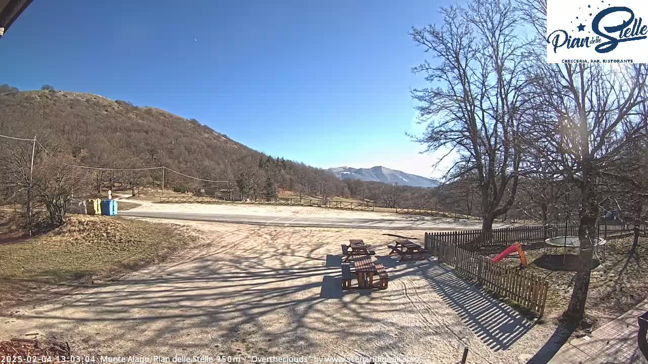 A sunny day reveals a mountainside parking area with picnic tables and a small playground, enclosed by a wooden fence.