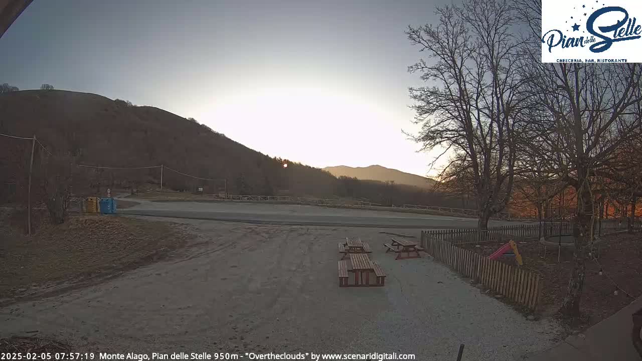 A sunrise illuminates a mountainside parking area with picnic tables and a small playground, enclosed by a wooden fence.