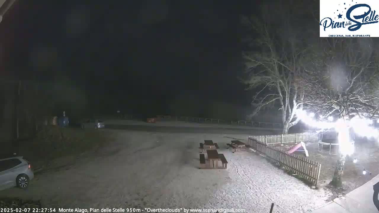 A snow-covered parking lot at night, with picnic tables, a small playground, and a few cars visible under dark, overcast skies.