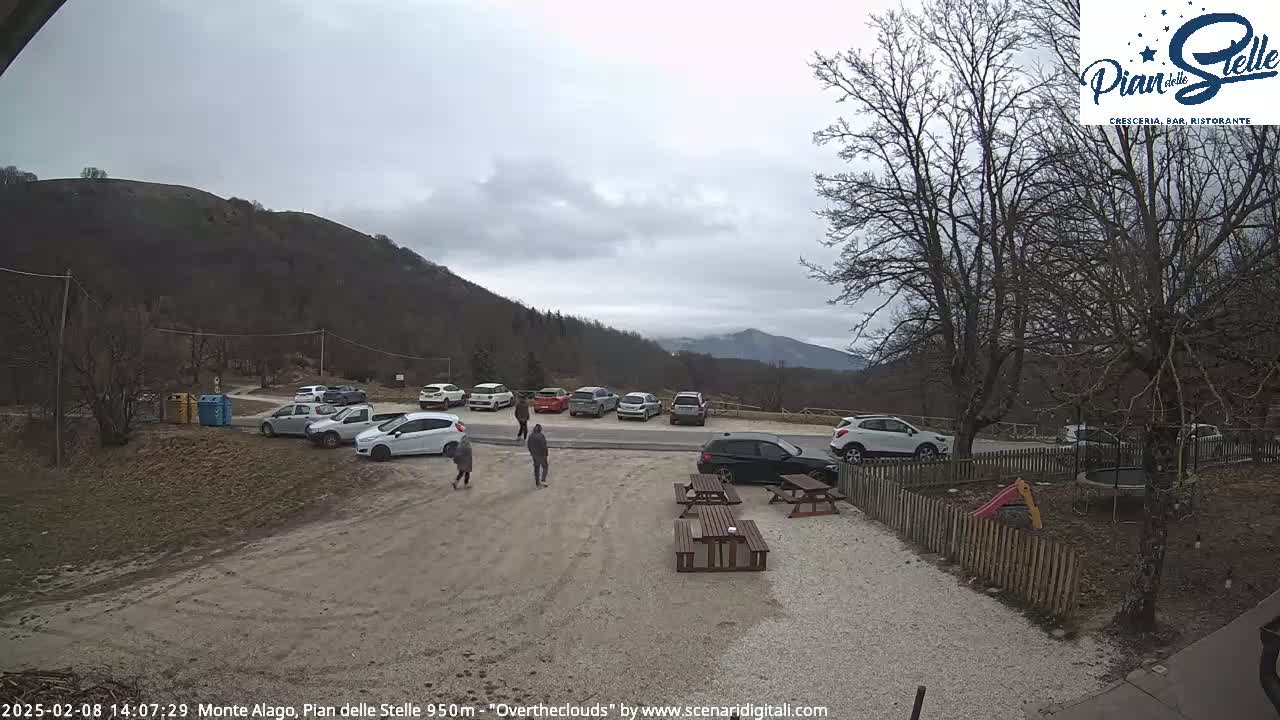 A parking lot with several cars is situated near a mountain range under an overcast sky.