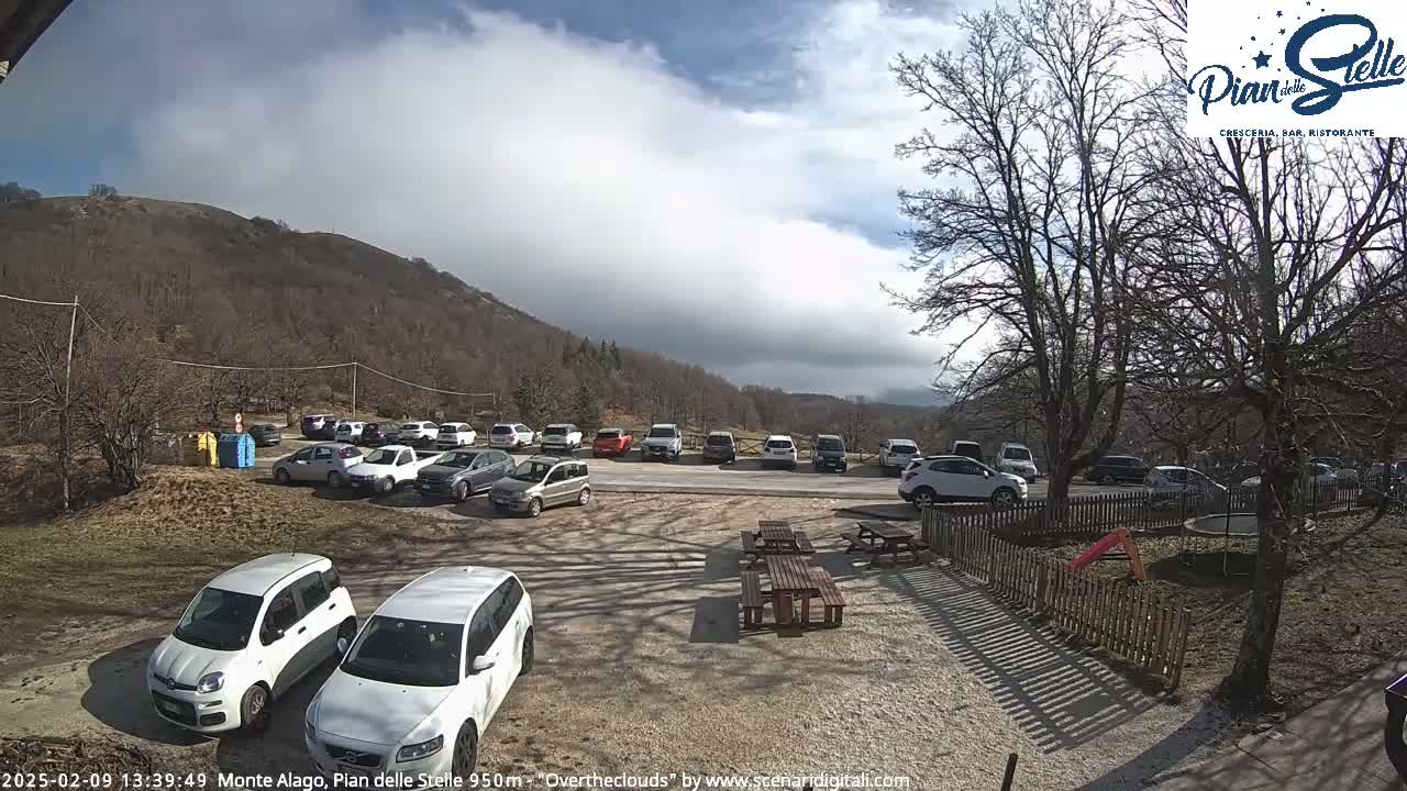 A parking lot filled with cars is situated near a mountainside under a partly cloudy sky.
