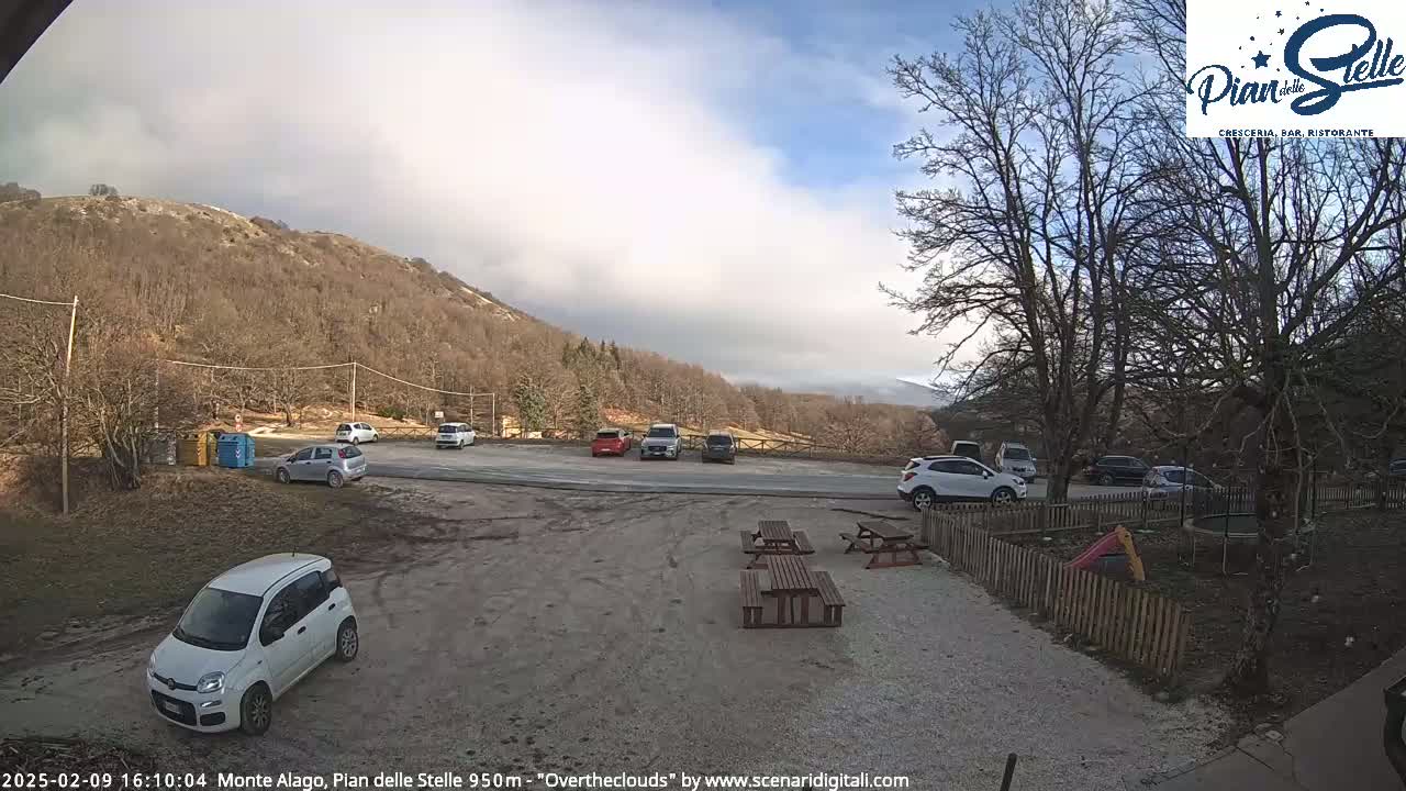 A parking lot with several cars is situated near a wooden fence and picnic tables, with a mountain in the background under a partly cloudy sky.