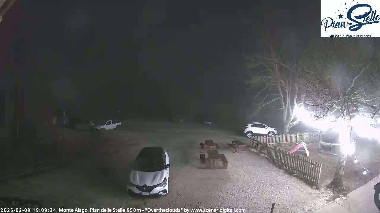At night, under an overcast sky, a gravel parking lot with several cars and picnic tables is illuminated, adjacent to a fenced yard with a trampoline and playground equipment.