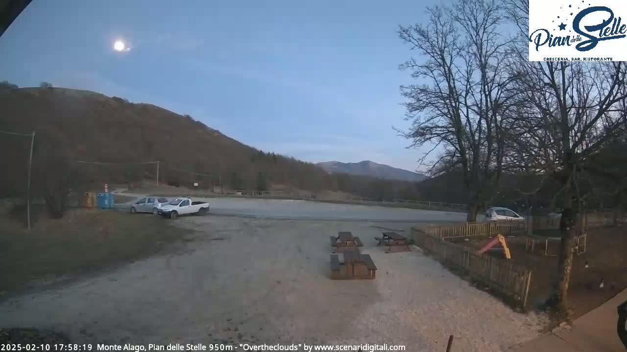 A mostly clear twilight sky with a bright moon hangs over a mountainside parking lot containing several vehicles, picnic tables, and a small playground.
