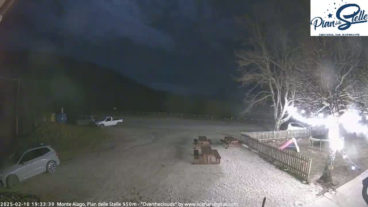 A nighttime outdoor scene shows a gravel area with picnic tables, a small playground, a few parked vehicles, and a tree with string lights under an overcast sky.