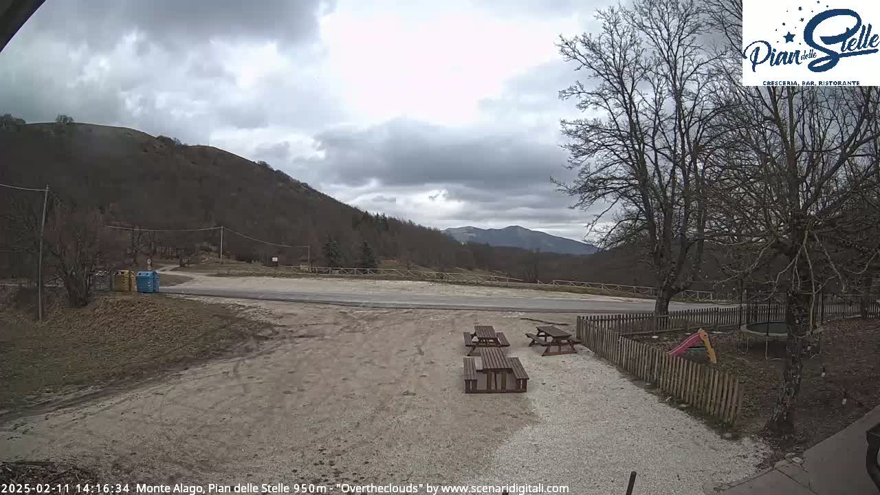 An overcast day reveals a mountain vista, a paved area with picnic tables, and a small playground enclosed by a fence.