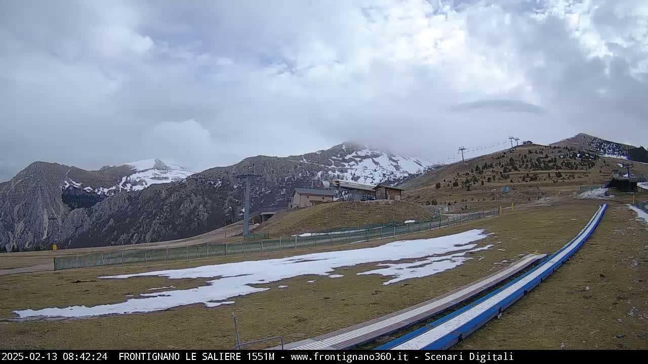 A mostly cloudy day reveals snow-dusted mountains in the background, with a ski lift and a few buildings visible on a grassy hillside speckled with patches of snow, and two long, blue ramps in the foreground.