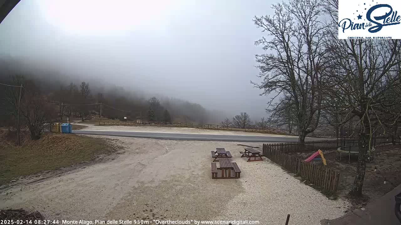 A foggy mountain vista overlooks a paved area with picnic tables and a small playground, enclosed by a wooden fence.