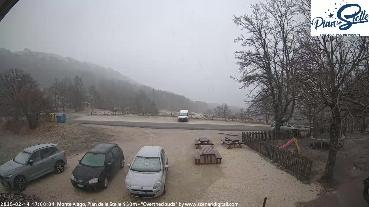 A snow-covered parking lot with several cars and picnic tables is seen in front of a mountain range under a heavy, gray, snowy sky.