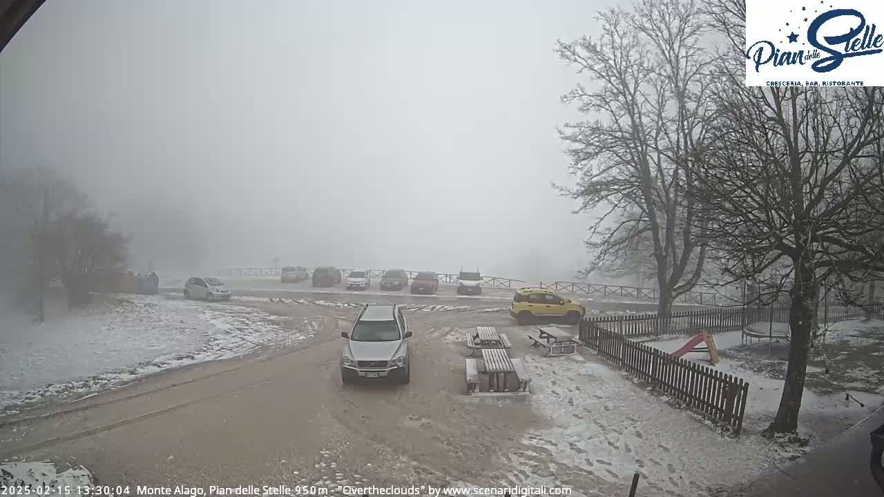A snowy parking lot with several cars, picnic tables, and a playground is shrouded in thick fog.