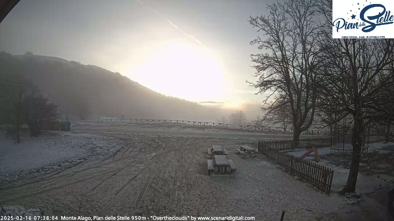A hazy sunrise illuminates a snow-covered landscape with picnic tables and a playground, nestled between a hill and leafless trees.