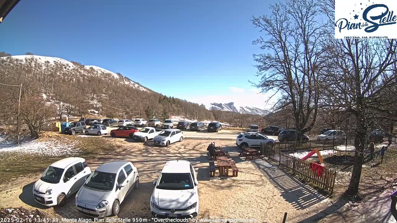 A sunny parking lot with numerous cars is nestled among snow-dusted, leafless trees and mountains under a clear blue sky.