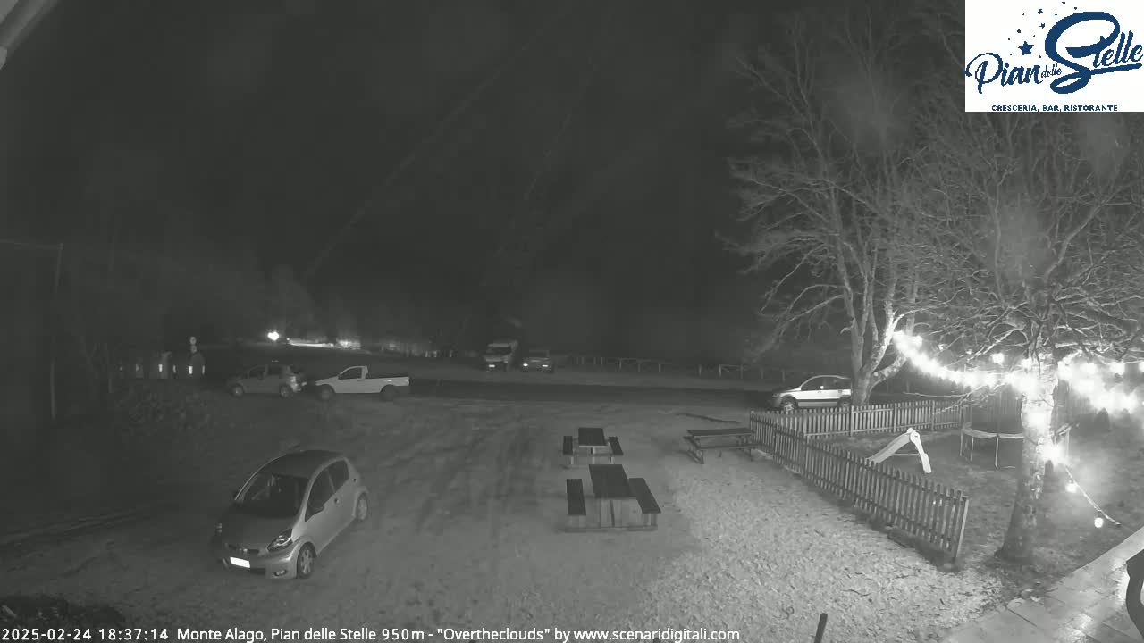 A nighttime, snow-covered parking lot with several cars, picnic tables, and a playground visible under an overcast sky.