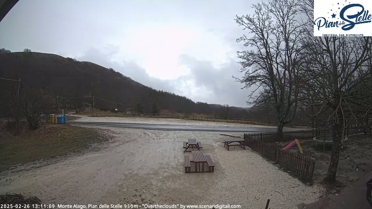 A muddy, overcast area with a few picnic tables near a wooden fence and bare trees, a road, and a hill in the background.