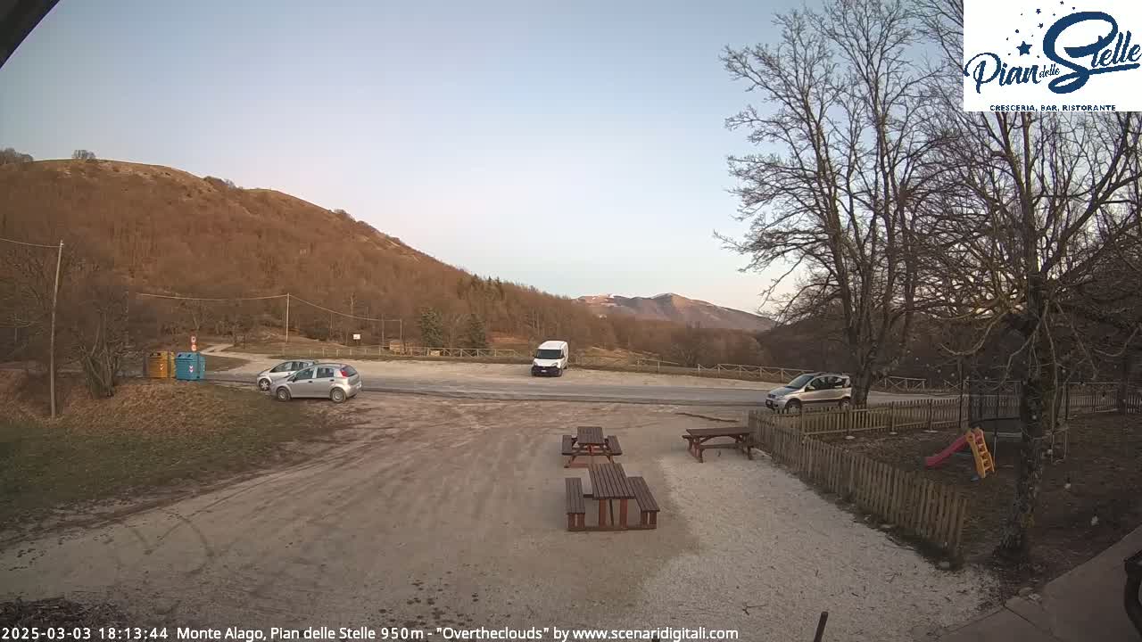 A mostly empty parking lot with a few cars and a van, picnic tables, and a playground is nestled between bare, brown hills under a clear, twilight sky.