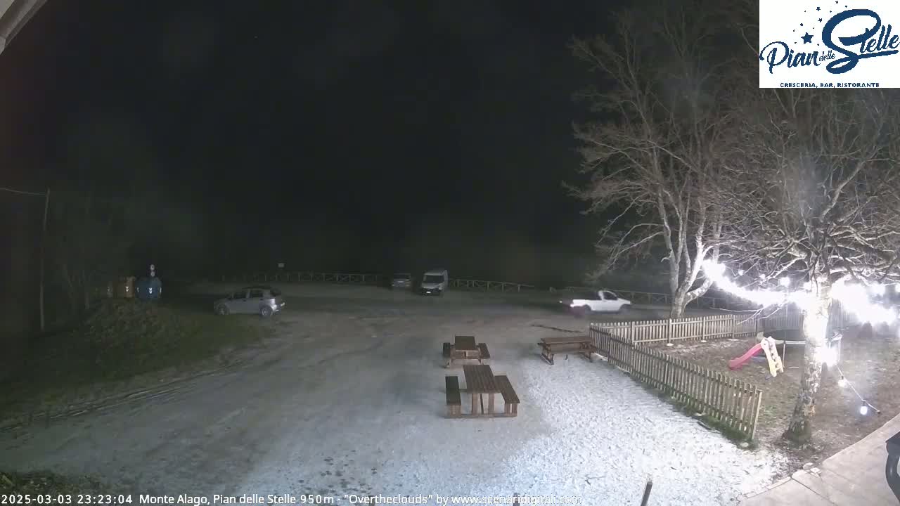 A snow-covered parking lot at night, with several vehicles parked, picnic tables, a small playground, and string lights strung around a tree.