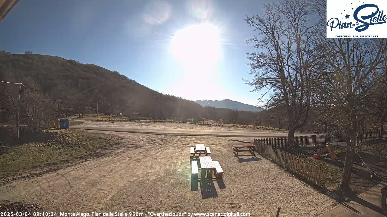 A sunny day reveals a gravel area with picnic tables, a wooden fence, and a playground, all set against the backdrop of a mountain range under a clear sky.